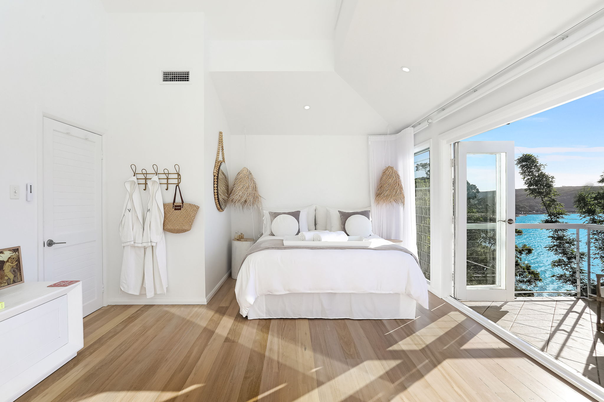 Bright white bedroom with robes, rattan pendant and open balcony doors to the ocean