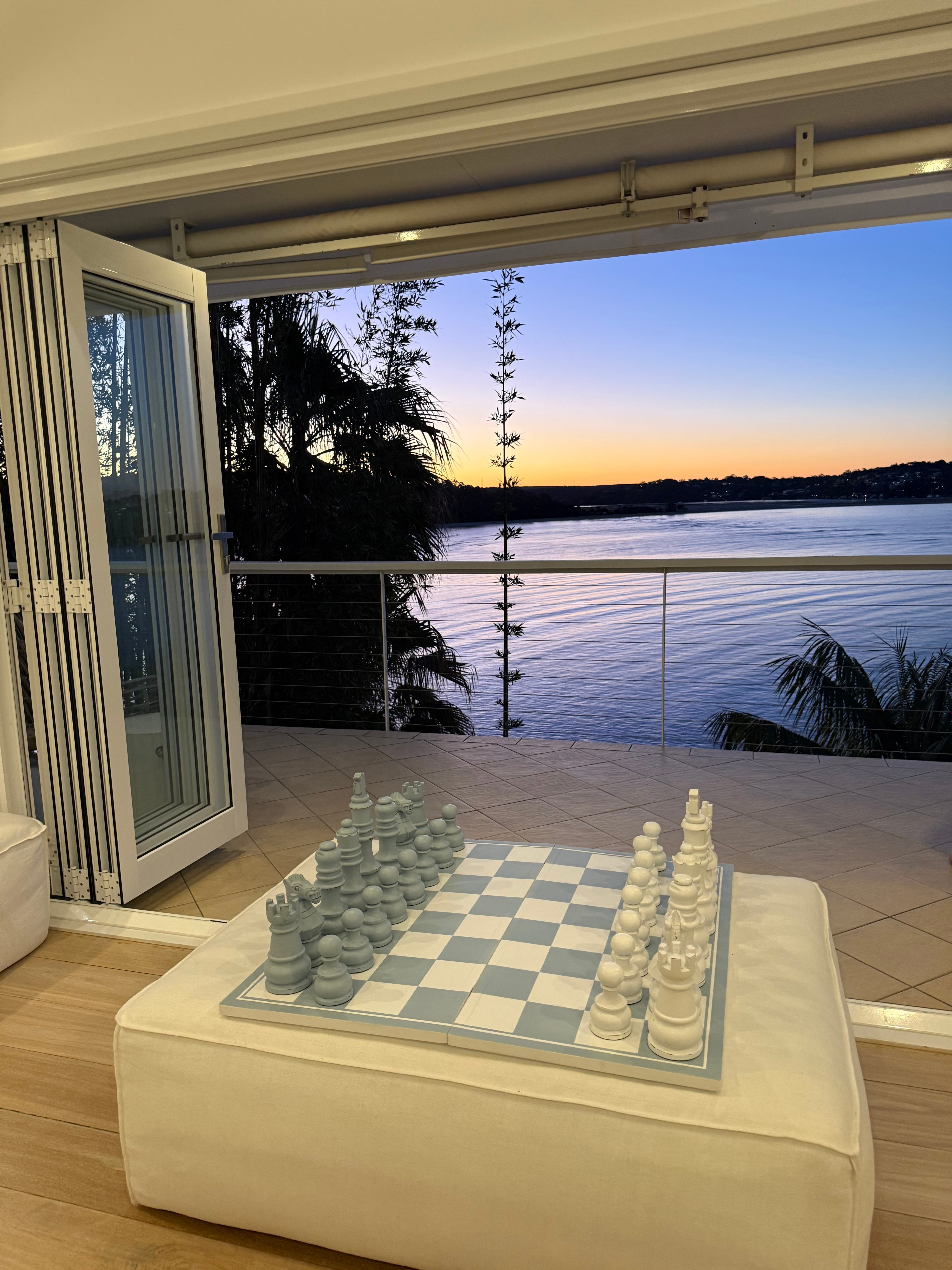 Chess set on white ottoman with vivid orange and blue twilight over the bay through open bifold doors